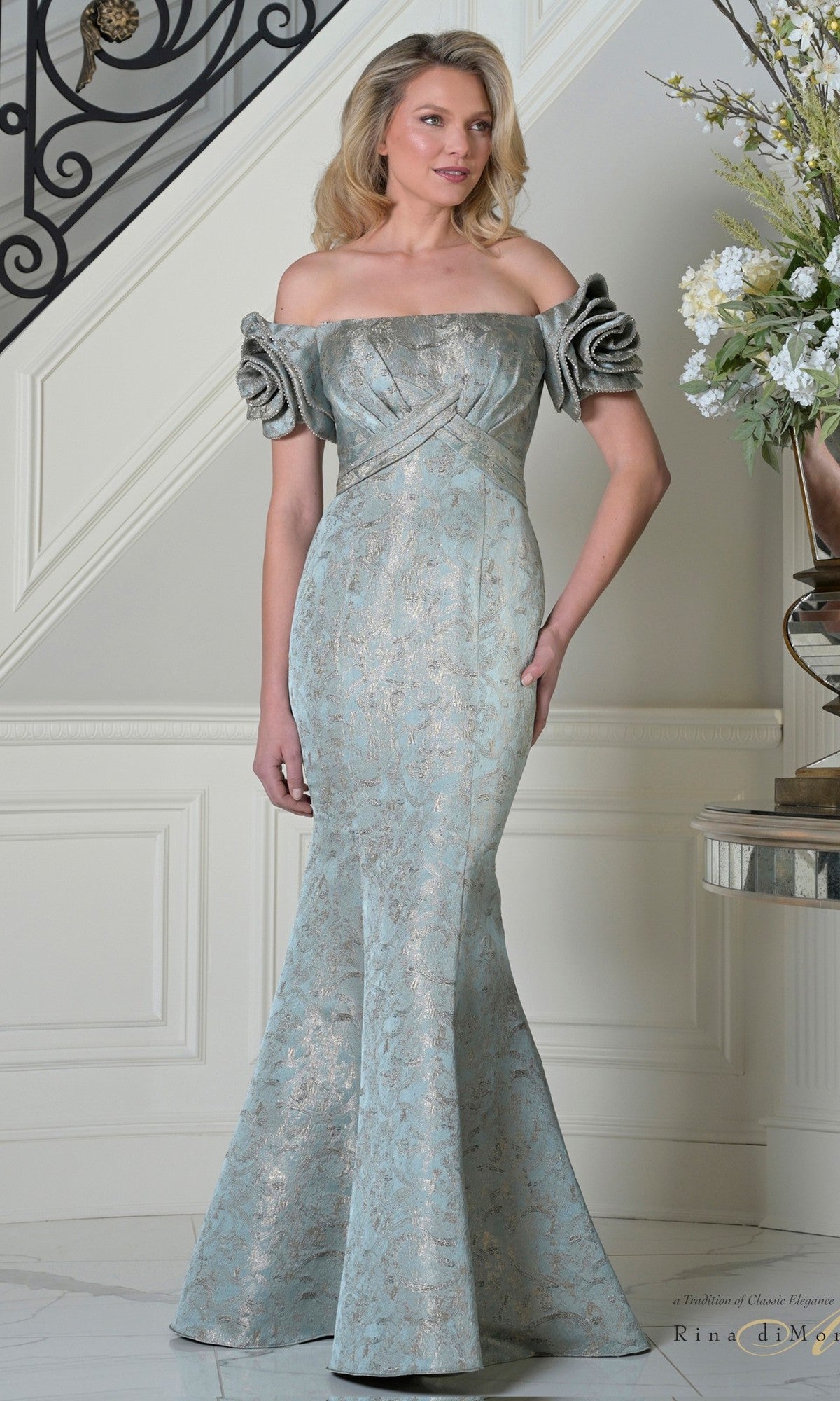A woman stands indoors wearing the Long Formal Dress: Rina Di Montella RD3512 in deep sage, a floor-length gown with a mermaid silhouette and dramatic off-shoulder rose sleeves.